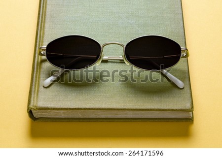 Summer reading items, an old book and sunglasses on yellow background.