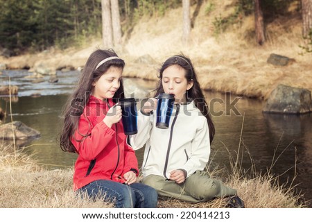 Two happy young girls resting beside a stream and talk with refreshing beverage