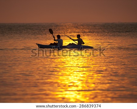 A couple in a double kayak paddle through the ocean, silhouetted by the setting sun on Koh Kood, Thailand.