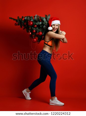 sexy Christmas fitness sport woman wearing santa hat holding xmas tree on her shoulders. standing on a red background. expression of emotions