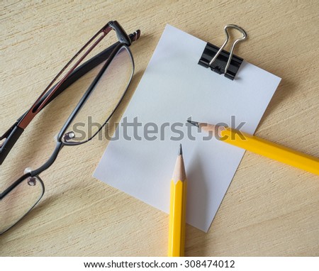 Blank white notepad, notebook, paper clips, yellow pencils and spectacles on vintage wood background