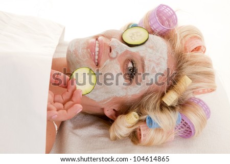 A woman has curlers in her hair and a cream face mask holding a cumber slice.