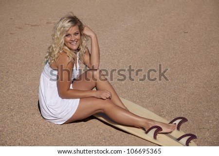 A woman sitting in the sand with her legs on her surf board with a smile on her face.