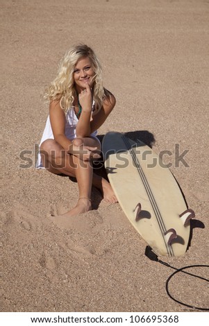 A woman sitting in the sand next to her surf board with a smile on her face.