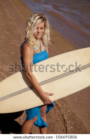 A woman holding on to her surf board walking with a small smile on her lips