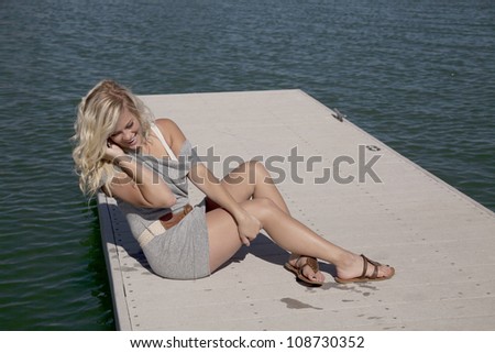 A woman is talking on the phone laughing, sitting on the dock.