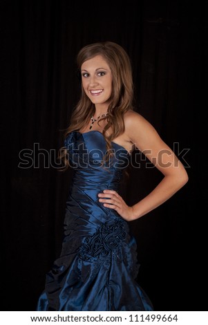 A teen with a blue formal dress standing in front of a black background with a smile on her face.