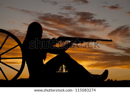 A woman sitting by a wagon wheel pointing her rifle .