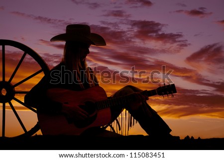 A cowgirl sitting up against a wagon wheel playing her guitar with the sunset behind her.