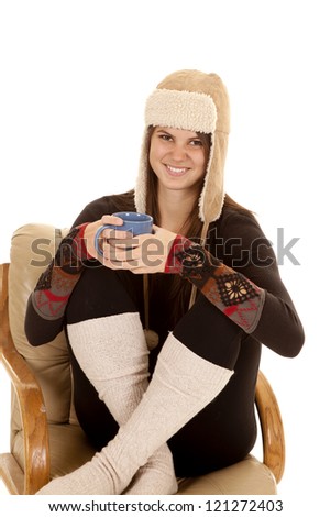 A woman sitting in her chair in her warm clothes and hat holding on to a mug with a warm drink.