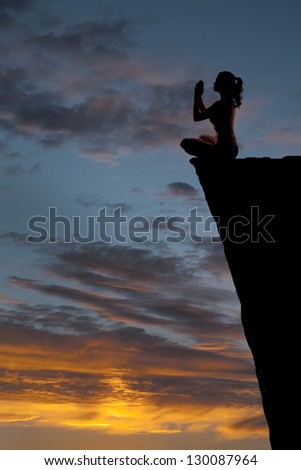 A woman is sitting on the edge of a high cliff in a prayer yoga position in the sunset.