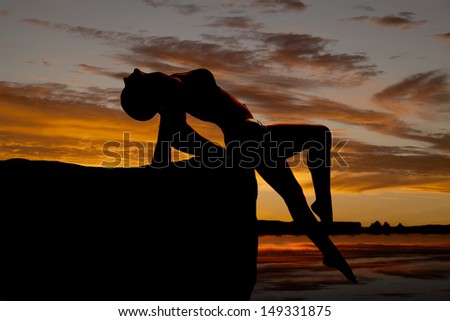 A silhouette of a  woman leaning back on a rock trying to reach the water
