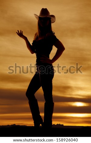 A silhouette of a woman in her jeans, boots, and hat standing in the outdoors with her hand up.
