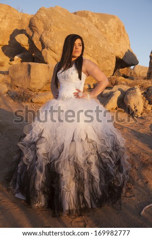 A woman in her formal dress standing in sand with rocks in the background.