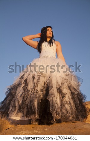 a woman in her formal dress standing on a rock with a beautiful blue sky.
