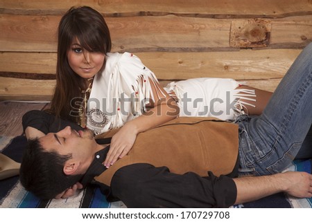A cowboy is laying on a blanket sleeping while an Indian is awake.