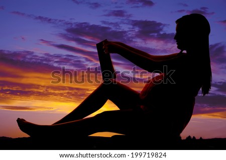 A silhouette of a woman sitting on the ground holding and reading a book.