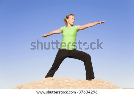 A woman on a rock with a blue sky doing the warrior two yoga move.