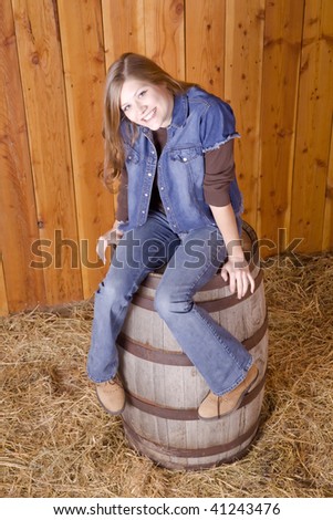 A woman sitting on a barrel in a barn with a wood wall background with a playful smile on her face.