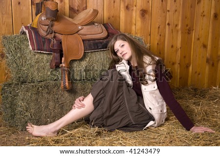 A woman sitting in the hay by a saddle with a skirt with her bare feet and legs showing with a serious look on her face.