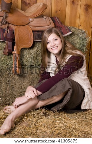 A woman sitting in the hay by a saddle with a skirt with her bare feet and legs showing with a smile on her face.