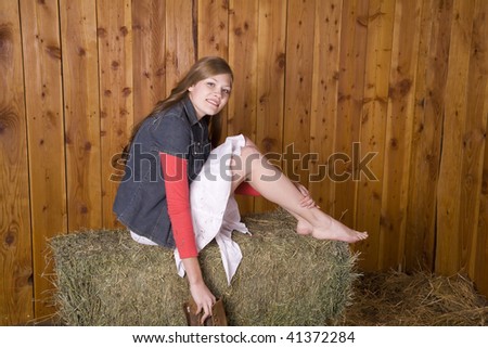 A woman sitting in a white skirt on top of a hay bale with bare feet holding her boots and smiling.
