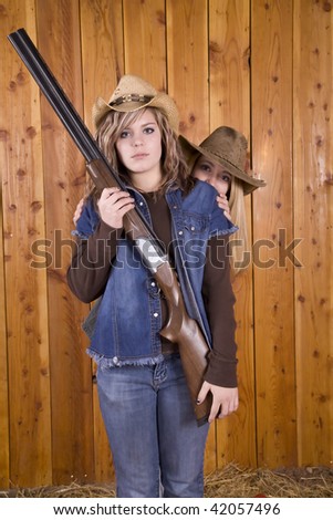 A girl standing with a shotgun with a serious look on her face with her friend standing behind hiding.