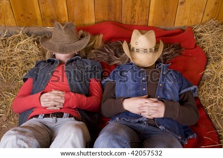Two girls laying on a red blanket in the hay with their hats over their faces covered.