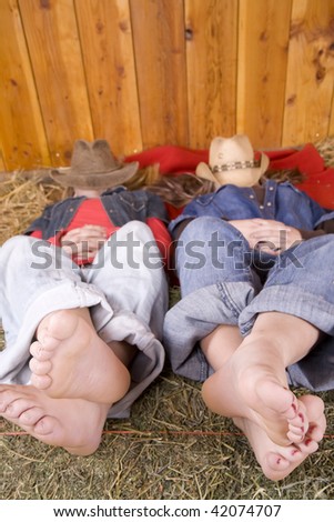 Two friends laying on a red blanket with bare feet up on a hay bale with hats on their faces relaxing.