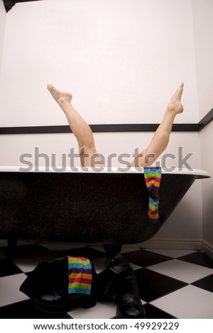 A woman in her tub playing around putting her legs up in the air in the splits, with her colorful striped socks hanging off the tub and her black boots on the floor.