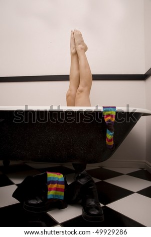 A woman in her tub relaxing with her legs up and a colorful sock hanging off the side of the tub and her black boots on her bathroom floor.