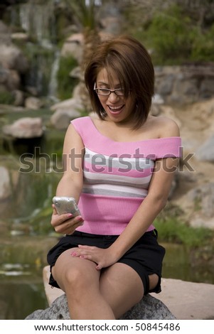 A woman sitting on a rock in a park by a pond getting a message with a surprised expression on her face.