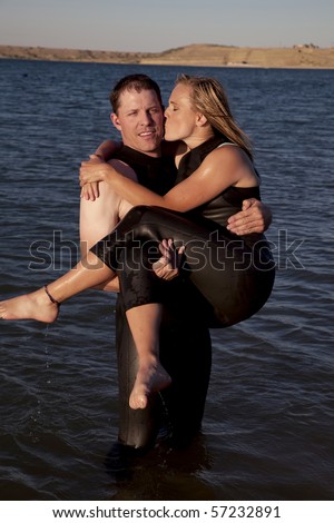 A couple together in their wet suits, showing their love by him holding her in his arms while she kisses him on the cheek.