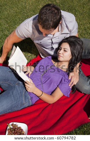 A couple is laying on a blanket reading a book.