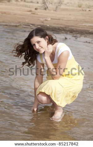 A woman kneeling down in her yellow dress using her finger to touch the water.