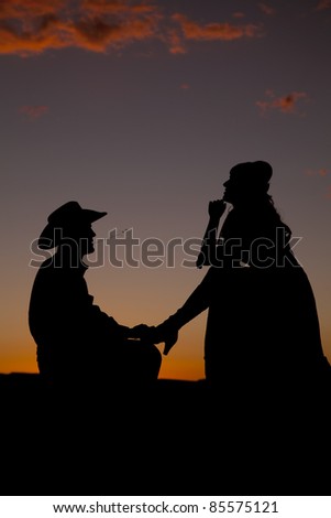 A cowboy is on his knees in the sunset with a woman by with one leg on him.