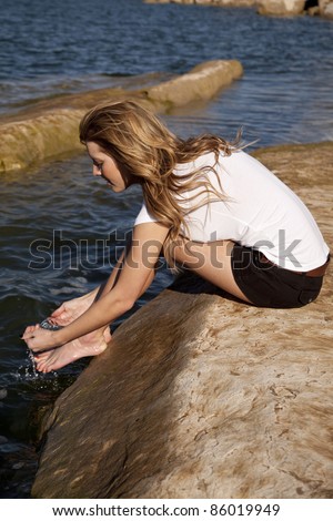 A woman sitting on a rock at the ocean putting water up on her feet to cool her off.