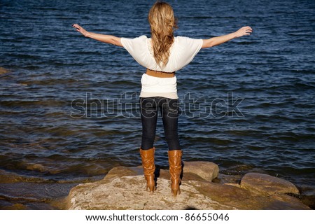 A woman with her back to the camera with her arms out looking over the water.