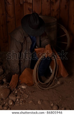 A cowboy kneeling down in the dirt looking down at the rocks.