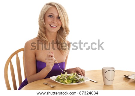 a woman sitting down in her fancy dress eating her salad with a smile on her face.
