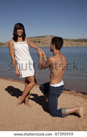 a man kneeling down and proposing to his woman on the beach with a shocked expression on her face.