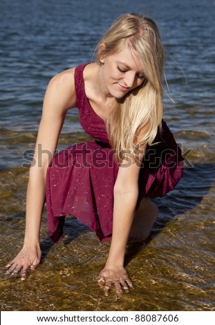 A woman kneeling down in her red formal with her hands in the water.