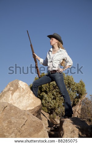 a woman standing up on top of rocks in her western wear holding a rifle.
