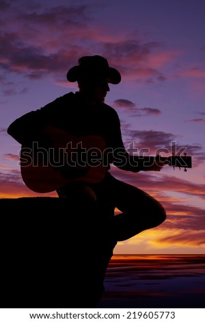 A silhouette of a man sitting on a rock ledge playing his guitar.