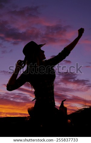 A silhouette of cowgirl sitting on her horse with her hand up in the air.
