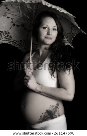 A black and white photo of a woman with a lace umbrella showing her baby belly and tattoo.