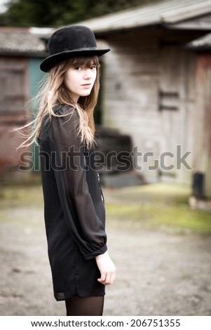 Portrait of a beautiful victorian style woman wearing a bowler hat and black dress standing in front of country farm stables