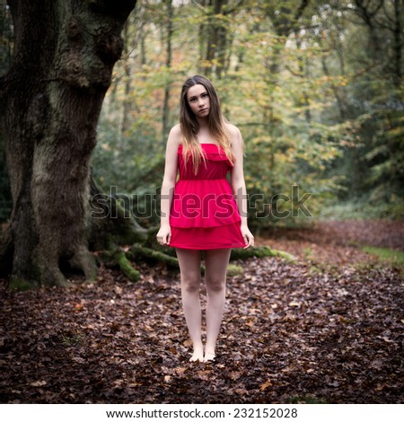 Portrait of a beautiful teenage girl with long dark blond hair wearing a short red dress having bare legs and feet standing in an autumn forest on a bed of leaves with an old tree in the background.