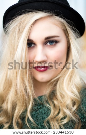 Portrait of a beautiful confident blond blue eyed teenage girl with long hair wearing a bowler hat, green t-shirt dark red lipstick standing alone in front of a fairground ride with a white fence.
