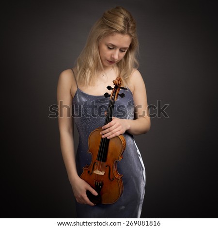 Studio portrait of a young woman with long blond hair in a grey and purple dress holding a violin looking down isolated against a dark grey background.